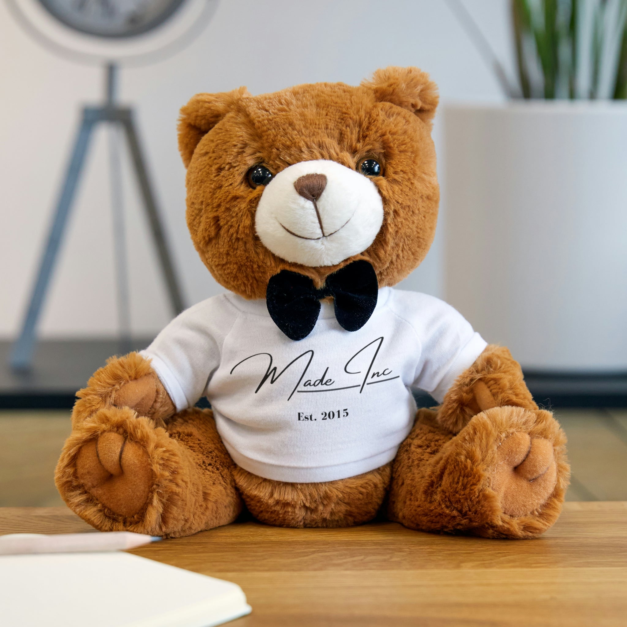 Image of a 7" by 10" teddy bear wearing a custom t-shirt, adorned with a charming bow. Made with luxuriously soft synthetic fur and 100% polyester filling, perfect for any celebration. brown plush teddy bear wearing a white Made Inc signature shirt and a black bow tie sitting on a hardwood floor in a studio